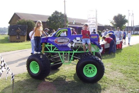 1995 Ford Bronco - A custom monster truck with a purple body and green chassis is displayed outdoors with people around.