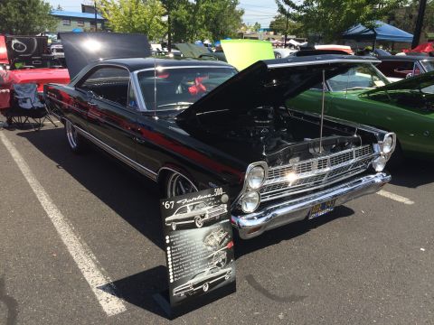 1962 Chevrolet Suburban - A black 1967 Ford Fairlane 500 is displayed with its hood open at an outdoor car show. A sign in front of the car provides details about it…