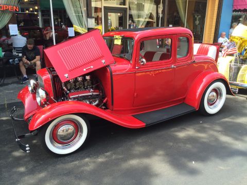 1962 Chevrolet Suburban - A bright red 1930s-style coupe hot rod with its hood open, revealing a V8 engine. The car is parked on asphalt in front of a storefront wit…
