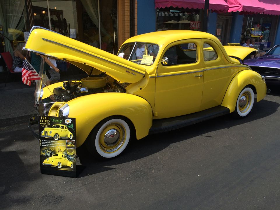 Yellow 1940 Ford Business Coupe with hood open, parked outdoors with a display sign.