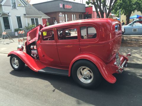 1962 Chevrolet Suburban - A red 1930s style hot rod sedan is parked on a street with its hood open, revealing the engine. A police car is visible in the background.