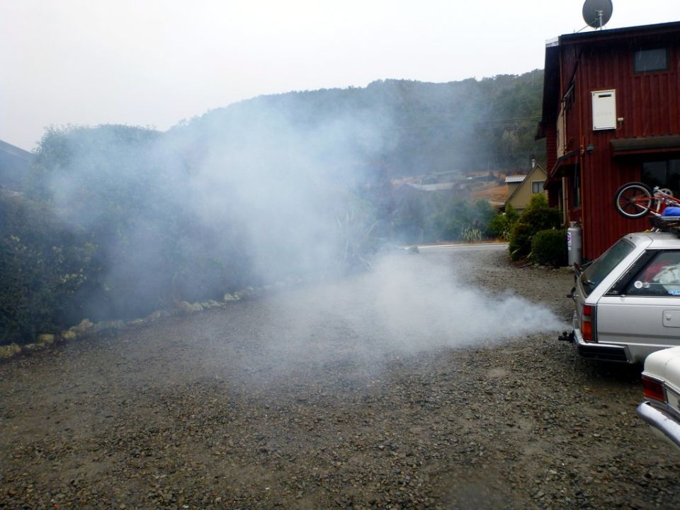 Silver station wagon emitting smoke from exhaust pipe, parked in front of a red building.