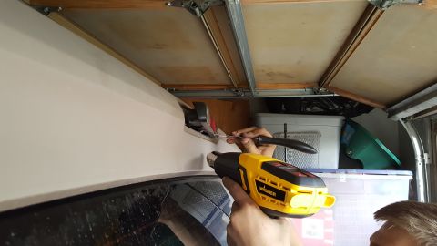 1996 Ford Bronco - A person uses a heat gun and a tool to apply trim to the top edge of a white vehicle, likely in a garage setting with storage bins and a ga…