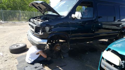 1996 Ford Bronco - A person works on the front wheel of a black van, with a spare tire and another vehicle nearby.