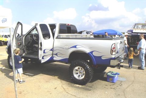 1995 Ford Bronco - A lifted pickup truck with custom flame decals is displayed with its doors open, while other vehicles and people are visible in the backgro…