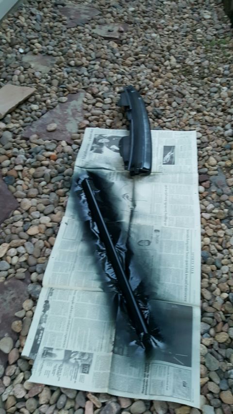 1996 Ford Bronco - A black trim piece is being spray painted on top of newspaper spread on a gravel surface.