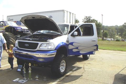 1995 Ford Bronco - Modified pickup truck with flame decals and trophies displayed at an event. A monster truck is visible in the background.