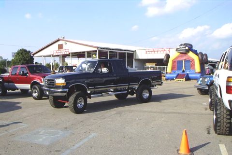 1995 Ford Bronco - Several lifted pickup trucks are parked outside a building with an inflatable attraction.
