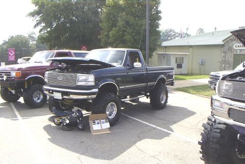 1995 Ford Bronco - Several lifted pickup trucks are parked in a lot, with one in the foreground having its hood open.