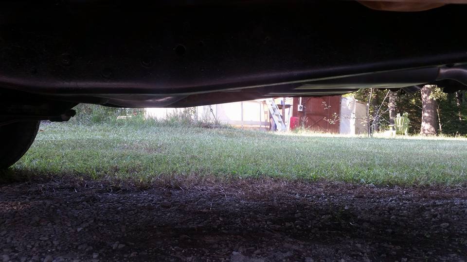 Underside view of a vehicle, showing the frame and suspension. Grass and trees in the background