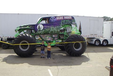 1995 Ford Bronco - The iconic Grave Digger monster truck is shown in a side profile, with two children posing in front of its massive tires.