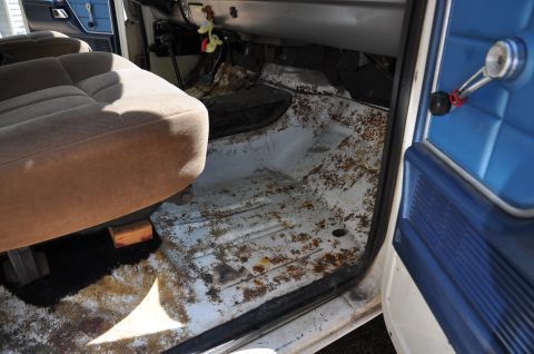 1979 Ford Bronco - The interior of a vehicle displays a worn seat, a heavily rusted floor, and a blue door panel with a chrome handle.
