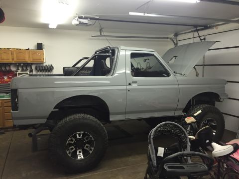 1988 Ford Bronco - Gray Ford Bronco with roll cage and lifted suspension sits in a garage with the hood open.