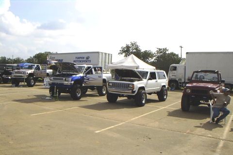 1995 Ford Bronco - A collection of modified trucks and SUVs are displayed outdoors under a partly cloudy sky.