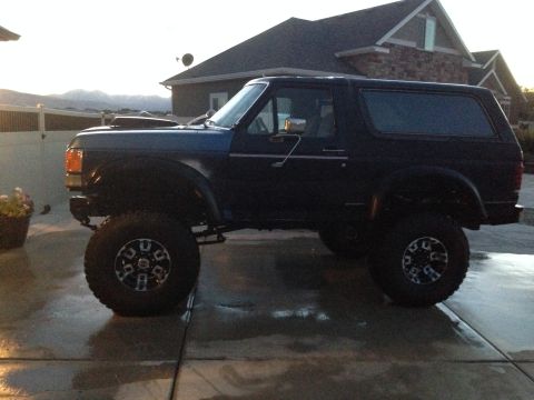 1988 Ford Bronco - A dark blue SUV with a lifted suspension and large tires is parked on a wet driveway.