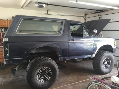 1988 Ford Bronco - This dark blue SUV is parked in a garage with its hood open, showing off its lifted suspension and aftermarket wheels.