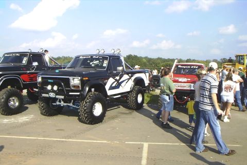 1995 Ford Bronco - Modified pickup trucks are displayed at an outdoor event, with several people milling around.