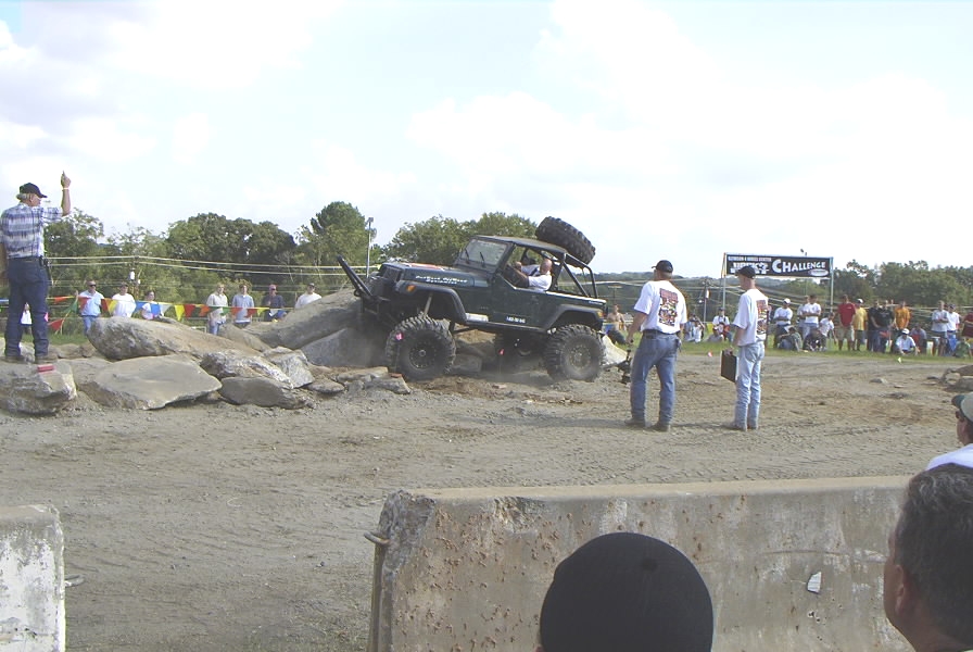 Green off-road vehicle with a spare tire on top, navigating a rocky obstacle course with spectators in the background