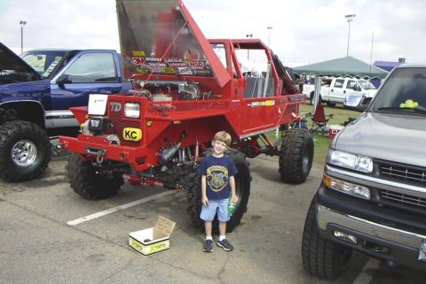 1995 Ford Bronco - A young boy poses in front of a heavily modified red off-road vehicle at an event, with other trucks in the background.