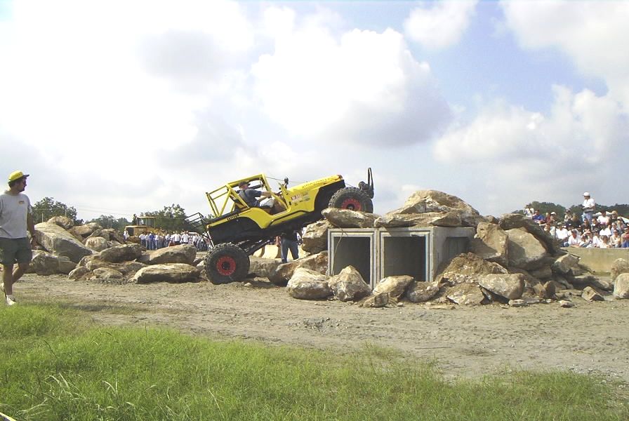 Yellow off-road vehicle with roll cage navigating a rock obstacle course, crowd watching in background