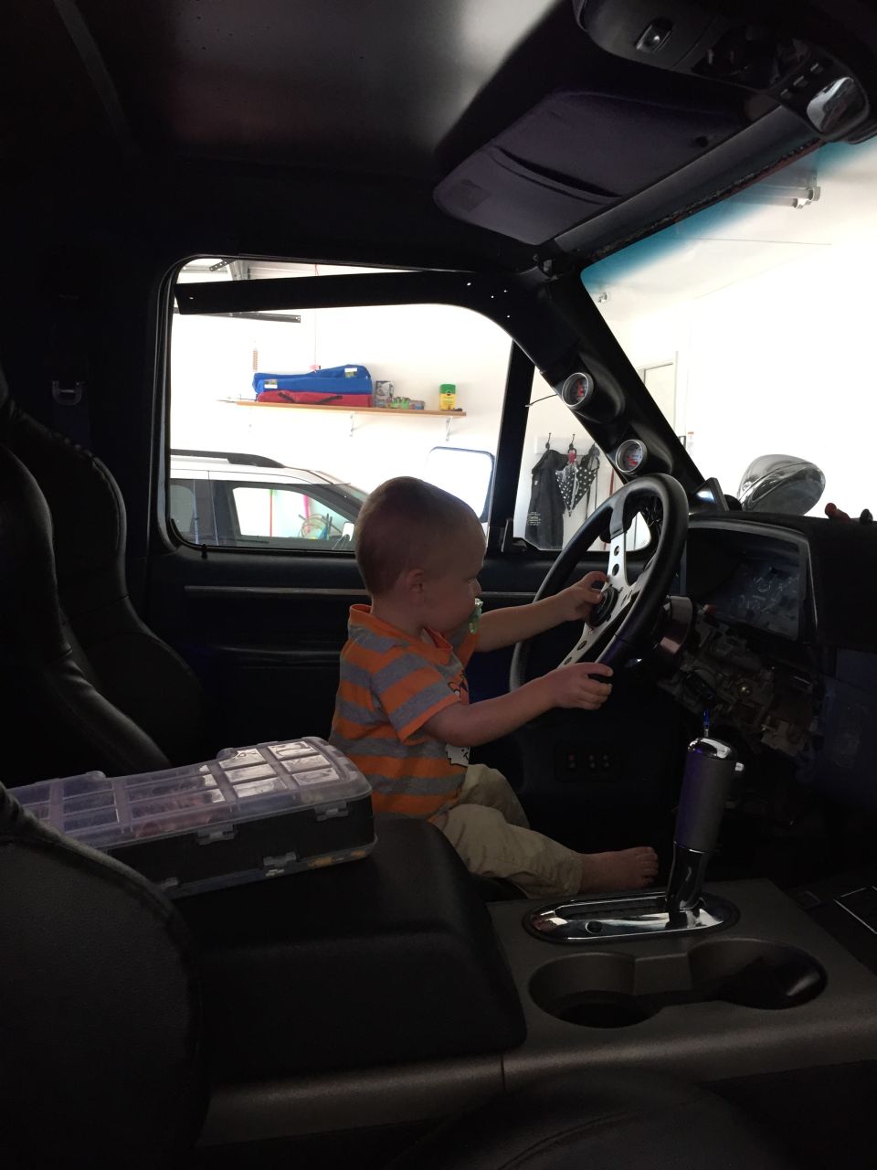 Child sits in driver's seat, hands on steering wheel. Toolbox on center console