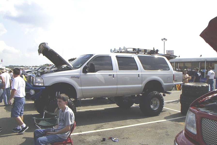 Lifted silver SUV, possibly a Ford Excursion, with hood open. Roof rack with lights. Parked at an outdoor event