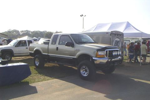 1995 Ford Bronco - A lifted gold and black Ford F-250 Super Duty pickup truck is parked next to a white truck and a trailer.