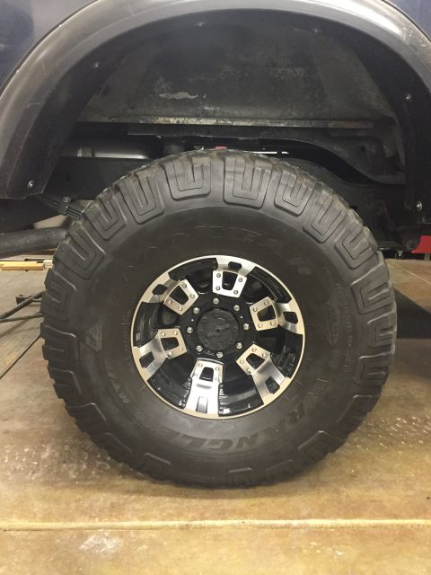 1988 Ford Bronco - A close-up, side view of a large, aggressive off-road tire and custom black and silver alloy wheel mounted on a vehicle. The tire tread is…