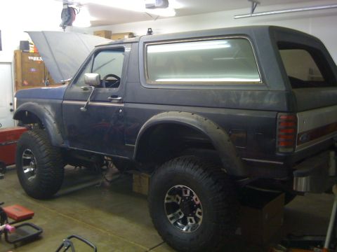 1988 Ford Bronco - A dark blue, lifted 1980s Ford Bronco is parked in a garage with its hood up. The vehicle has large off-road tires and fender flares.