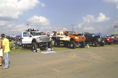 1995 Ford Bronco - Multiple custom lifted trucks displayed at an outdoor automotive event under a cloudy sky.