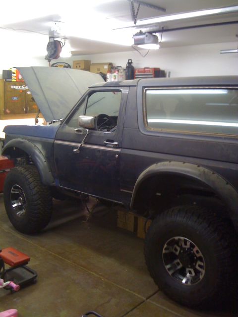 1988 Ford Bronco - A dark blue Ford Bronco is parked in a garage with its hood open. The vehicle has large off-road tires and aftermarket fender flares.