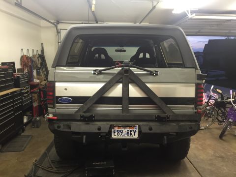 1988 Ford Bronco - The rear view of a silver Ford Bronco is shown in a garage. It features a custom rear bumper with a spare tire carrier mounted on the tailg…
