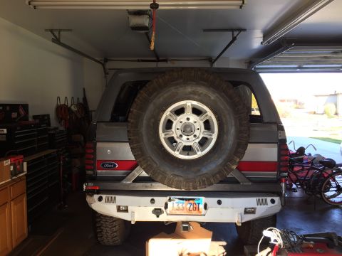 1988 Ford Bronco - Rear view of a gray Ford Bronco parked in a garage with a large spare tire mounted on the back. The vehicle has a custom rear bumper and a…