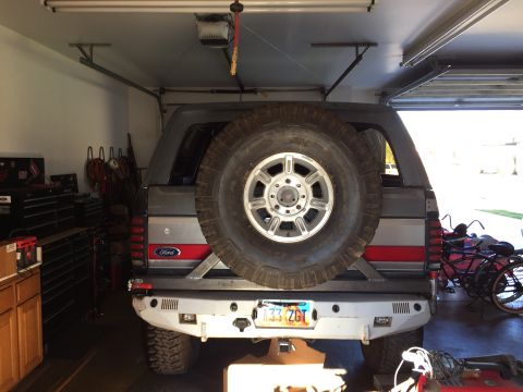 1988 Ford Bronco - Rear view of a lifted Ford Bronco parked in a garage with a spare tire mounted on the back. The vehicle has a custom rear bumper and a lice…