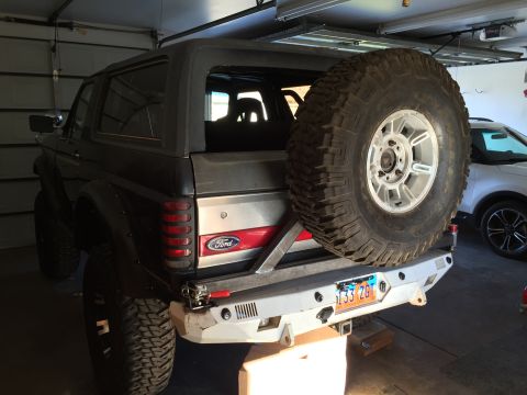 1988 Ford Bronco - The rear view of a dark-colored, lifted Ford Bronco is shown parked in a garage. It features a large spare tire mounted on the rear bumper,…