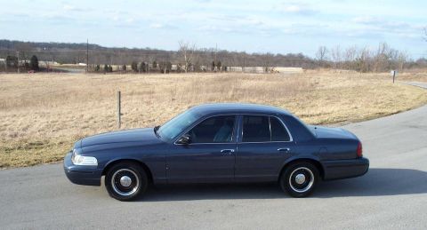 1998 Ford Crown Victoria - A dark blue 1998 Ford Crown Victoria sedan is parked on the side of a road with a dry field in the background.