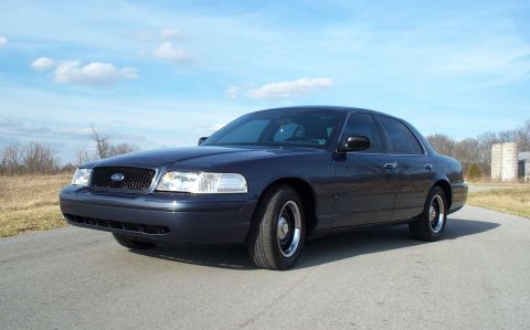 1998 Ford Crown Victoria - A dark blue 1998 Ford Crown Victoria is parked on a paved road under a partly cloudy sky.