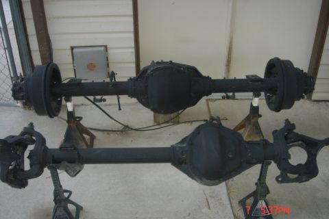 1988 Ford Bronco - Two black painted rear axles are displayed on jack stands outdoors. The upper axle has drum brakes, while the lower axle has visible steeri…