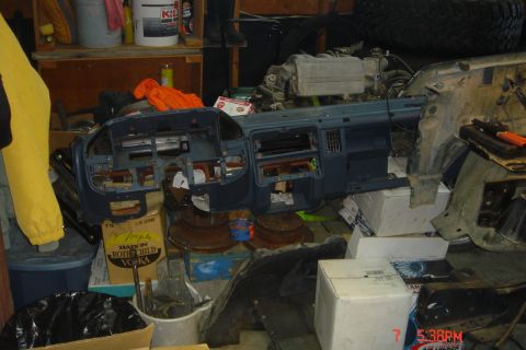 1988 Ford Bronco - A disassembled blue dashboard is laid out on the floor amidst various car parts and boxes in a garage setting. A yellow garment is draped o…
