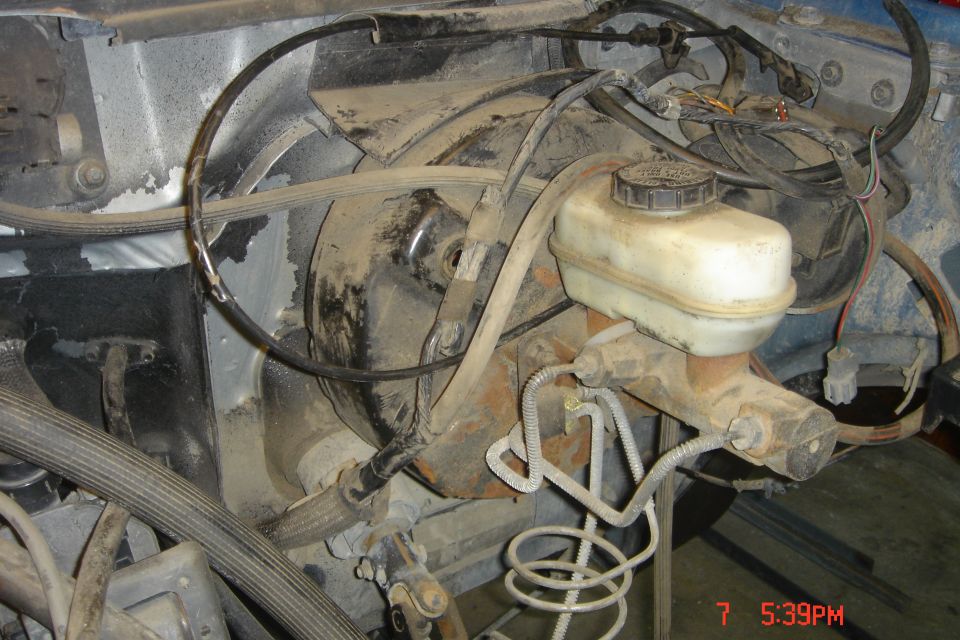 Close-up of dusty brake master cylinder and booster assembly in a vehicle engine bay.