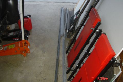 1988 Ford Bronco - A red creeper seat is leaning against a wall next to some metal framing. A floor jack and a red gas can are visible on the concrete floor.