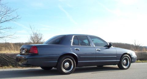 1998 Ford Crown Victoria - A blue Ford Crown Victoria is parked on the side of a road with a wooded area in the background.