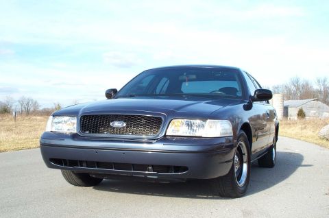 1998 Ford Crown Victoria - A dark blue 1998 Ford Crown Victoria is parked on a paved road with a rural background under a clear sky.