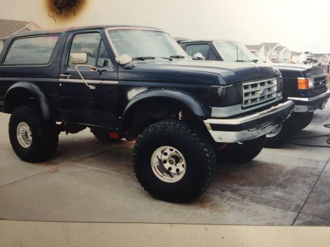 1988 Ford Bronco - A dark blue Ford Bronco is parked on a concrete driveway next to a similar model truck. The Bronco has large off-road tires and appears to…