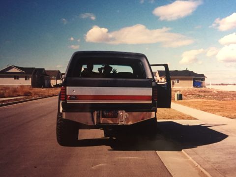 1988 Ford Bronco - Rear view of a dark blue Ford Bronco parked on a suburban street. The vehicle has a red and white stripe across the tailgate and an open dr…
