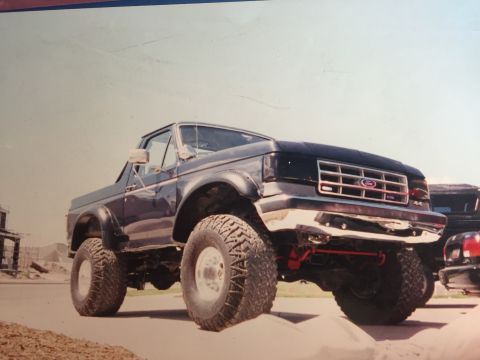1988 Ford Bronco - A lifted, dark blue 1988 Ford Bronco is shown from a low angle, showcasing its large off-road tires and chrome bumper. The vehicle is parke…