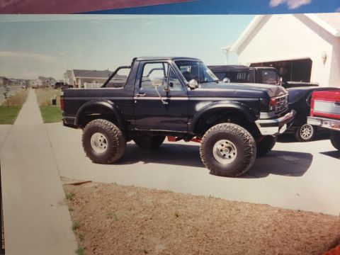 1988 Ford Bronco - A dark blue Ford Bronco with large off-road tires is parked on a paved driveway in front of a white house. The vehicle is modified with a r…