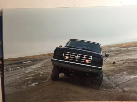 1988 Ford Bronco - A black Ford Bronco is shown from a low front 3/4 angle driving on a wet, muddy, and sandy terrain under an overcast sky. The vehicle has a…