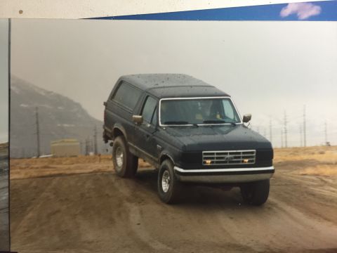 1988 Ford Bronco - A dark-colored Ford Bronco is shown from a three-quarter front angle, driving on a dirt road with a mountainous, overcast landscape in the…