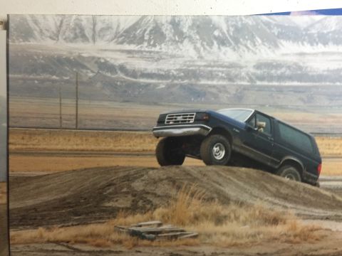 1988 Ford Bronco - A dark blue Ford Bronco is captured mid-air, driving up a dirt mound with snow-capped mountains in the background.